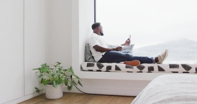 Adult African American man sitting in window nook checking smartphone while relaxing