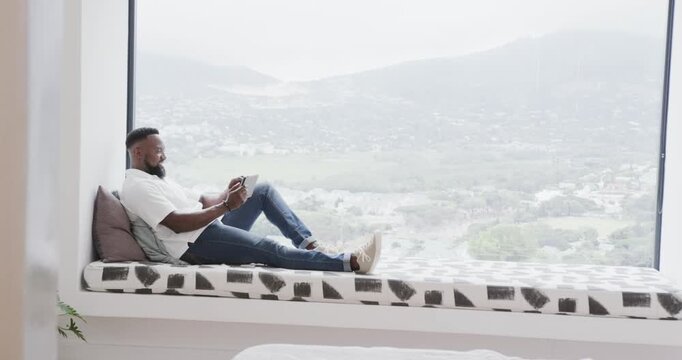 Man settling into window bench, reclining by large window and reading tablet to relax, copy space