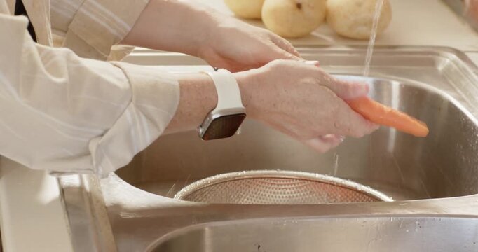 Woman washing carrot under running faucet at kitchen sink, rinsing in colander, wearing striped top