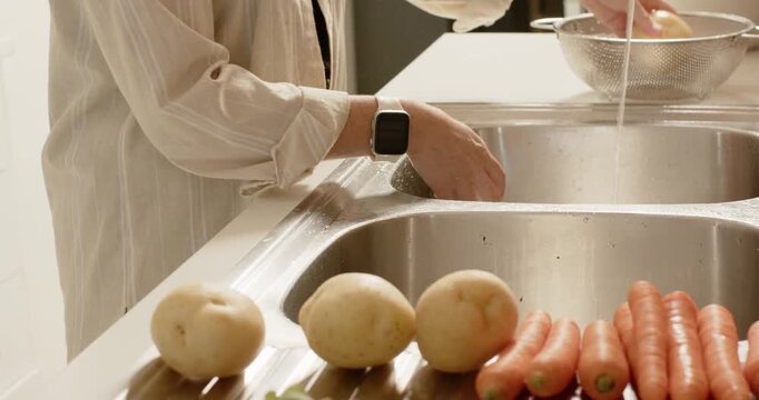 Woman in striped shirt turning on faucet, rubbing and rinsing potatoes into colander for cooking