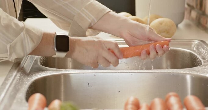 Woman's hands reaching and rinsing carrot under running faucet in sink with potatoes, prepping meal