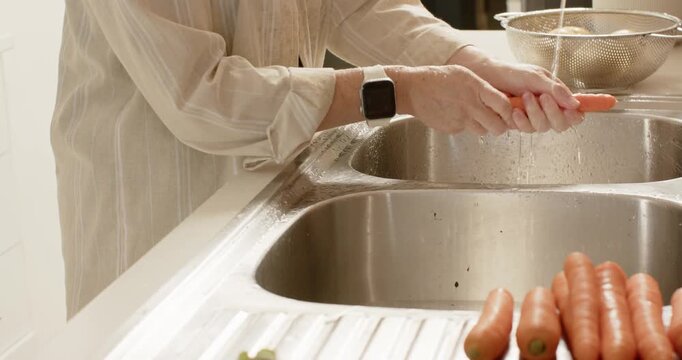 Adult female wearing blouse washing carrots at sink with running faucet into colander for meal prep