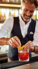Male bartender in a black vest prepares a cocktail by adding a lemon twist to a glass on the bar counter with bottles and glasses in the background
