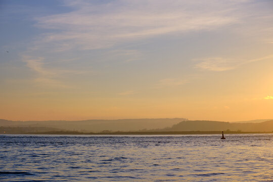 March 21 2026. A serene sunset seascape from Exmouth, a lone navigation buoy on the golden waters of the Exe estuary, hazy East Devon hills fading into the orange horizon.
