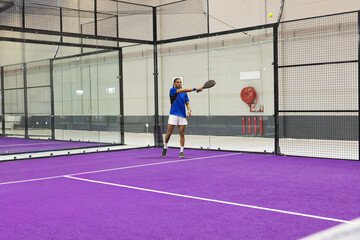 Obraz premium Man playing paddle on purple turf court, wearing blue shirt and white shorts, hitting yellow ball