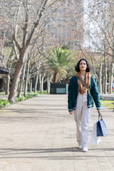 Obraz premium Woman walking down paved tree-lined path carrying shopping bags wearing scarf and jacket