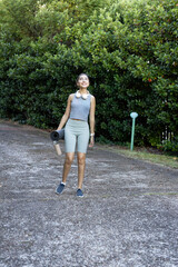 Fototapeta premium Indian woman walking on paved path by hedge in activewear carrying yoga mat, bottle, headphones