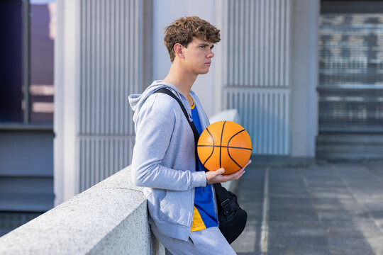 Teen male leaning on concrete railing on rooftop, holding orange basketball, in hoodie with bag