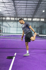Obraz premium Adult male athlete balancing and stretching quads on purple synthetic court by net, paddle nearby