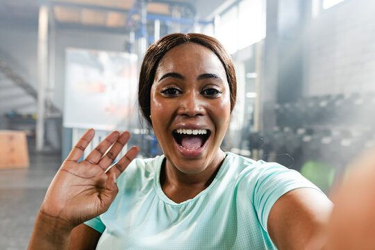 Mid-adult African American woman waving and taking selfie at gym in mint shirt near dumbbells