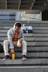 African American man sitting on steps at plaza with headphones, holding sandwich, grabbing juice © wavebreak3