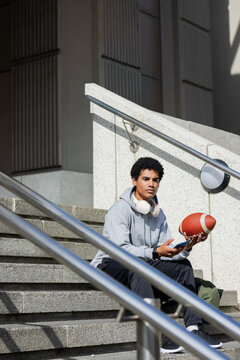 African American man sitting on campus steps holding football smartphone and headphones
