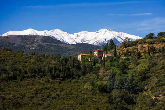 paysage de montagnes enneig&eacute;es sous un ciel clair, avec des sommets imposants en arri&egrave;re-plan. Au premier plan, on v&eacute;g&eacute;tation dense de conif&egrave;reset des b&acirc;timents traditionnels aux toits rouges.