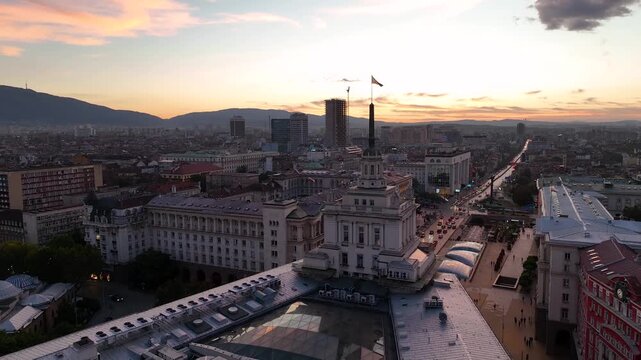 A close-up view of the flag of Bulgaria blowing in the wind against a sunset or sunrise over the capital city of Sofia. The Bulgarian flag flying over the Parliament building.