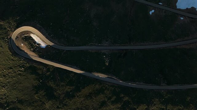 Top down Aerial drone footage of dramatic serpentine curves of Beartooth Highway cutting through dark volcanic alpine terrain with patches of snow and ice scattered across rugged mountain landscape.