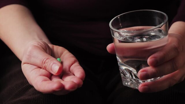 A close-up of hands holding pills and a glass of water. A person is preparing to take medication. Conceptually related to healthcare, treatment, illness, vitamins, or pain relief.