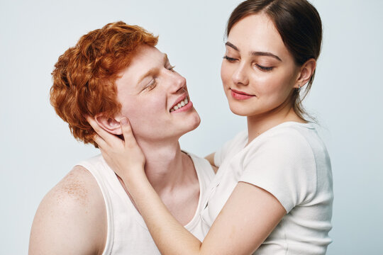 Couple in casual white clothing embracing and smiling with affection on light background. Young woman gently holds red-haired man showing intimacy and connection indoors.