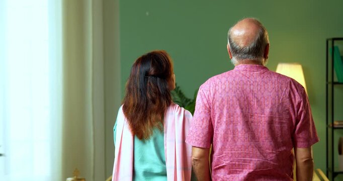 A back-view shot of a senior Indian couple standing in their living room, pointing toward the distance. The retired husband and wife share a moment of planning or admiration at home