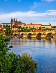 Obraz premium A vibrant scenic view of a historical stone bridge, castle, and waterway under a clear, bright blue sky with fluffy white clouds
