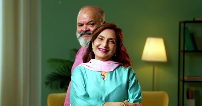 A senior Indian woman uses her hands to create a "frame" gesture, visualizing a design or memory, while her husband watches with interest. A creative moment in their living room