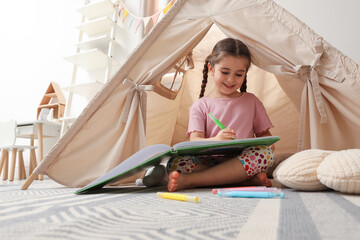 Little girl drawing in sketchbook near toy wigwam at home, low angle view © New Africa
