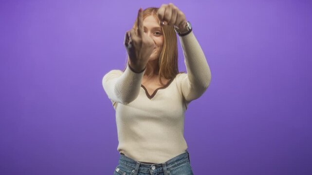 Woman shows middle finger to camera while pointing with other hand, wearing cream ribbed sweater and denim jeans against a purple studio backdrop; defiance attitude.