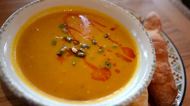 Bowl of creamy pumpkin soup topped with seeds and oil, served with lemon and bread