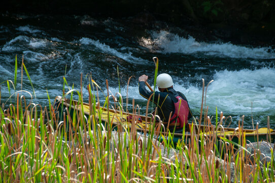 A lone canoer paddles upstream on fast river rapids near marshy reeds while wearing a helmet and life jacket. The scene evokes adventure, motion, outdoor recreation and rugged nature.