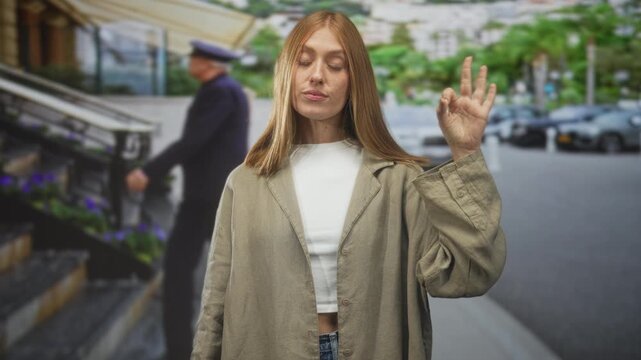 Woman redhead showing ok hand gesture with visible hand at building entrance on street near steps and parked cars; casual confidence.