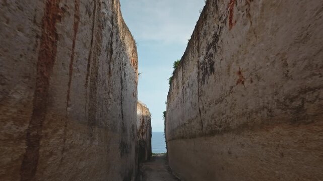 Towering limestone cliffs framing glimpse of tranquil Indian Ocean and sand beach below under bright blue sky. Pathway road winding through a deep limestone canyon in Bali, Indonesia. Drone flight