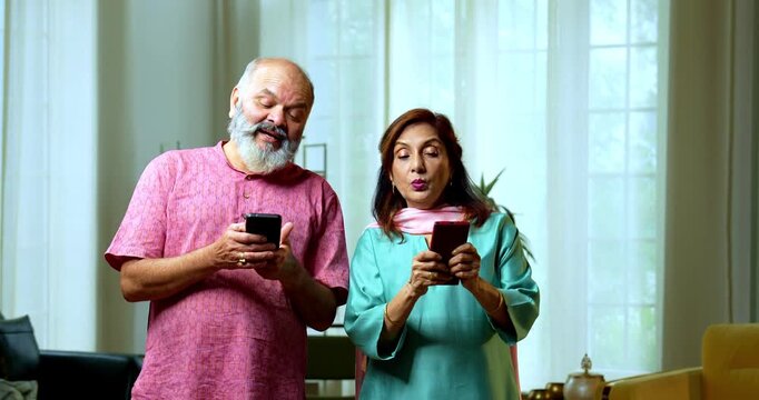 An Indian senior couple standing side by side and typing on their smartphones. They are playfully peeping into each other screens with curious and funny expressions in their living room