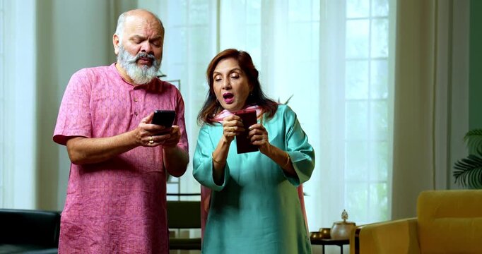 An Indian senior couple standing side by side and typing on their smartphones. They are playfully peeping into each other screens with curious and funny expressions in their living room