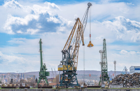 Industrial harbor scene featuring massive portal cranes against a cloudy sky, with a scrap metal pile and coastal hills in the background. Port infrastructure photography