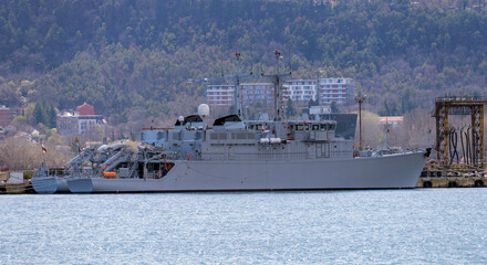 Large grey military vessel docked at a port with a backdrop of a forested hillside and coastal buildings. The warship features radar masts, specialized equipment, and a sleek naval hull © John_Doo78
