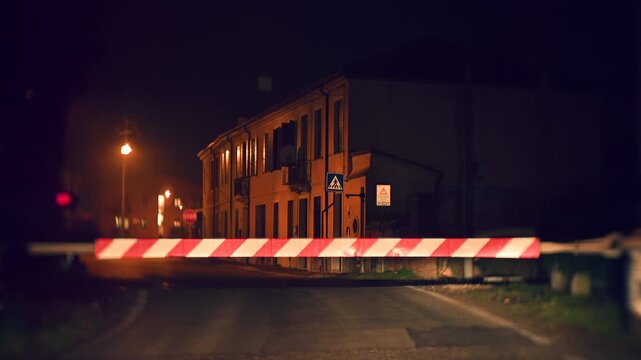 Night video of a closed railway crossing in a village street with houses nearby. Warning lights active while a train covered with graffiti passes through. Atmospheric scene combining transport