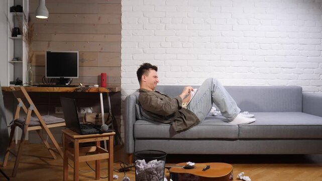 Young man relaxing on the couch and looking at his smartphone, avoiding work on the laptop nearby, surrounded by a messy room and crumpled papers, showing a scene of procrastination and idleness
