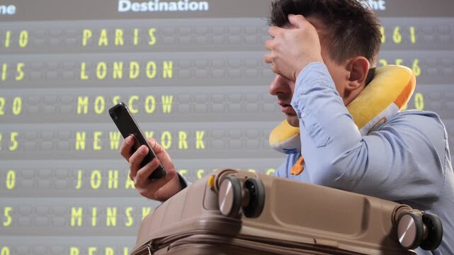 Stressed man leaning on his suitcase, holding a smartphone, looking frustrated and tired in front of a flight departures board, experiencing travel delays at a busy airport