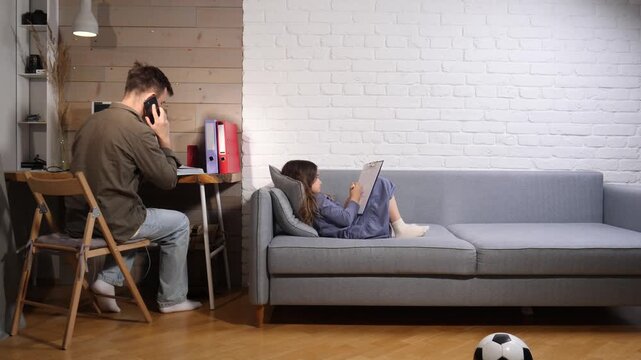 Father working from home at a desk, talking on a smartphone while daughter relaxes on the sofa drawing nearby modern family multitasking, remote work and parenting balance