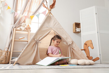 Little girl drawing in sketchbook near toy wigwam at home © New Africa