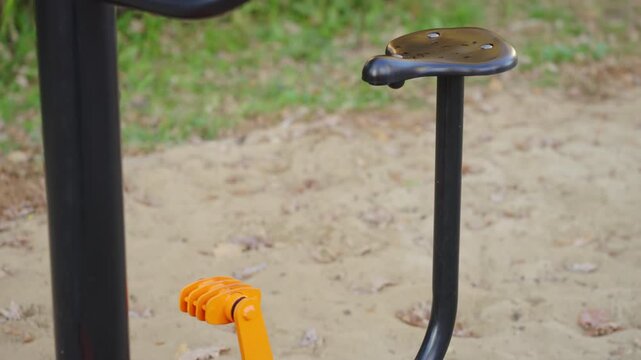 Fitness machine pedals curve around black post on sandy ground. Textured footrests support stepping motion marking outdoor workout zone