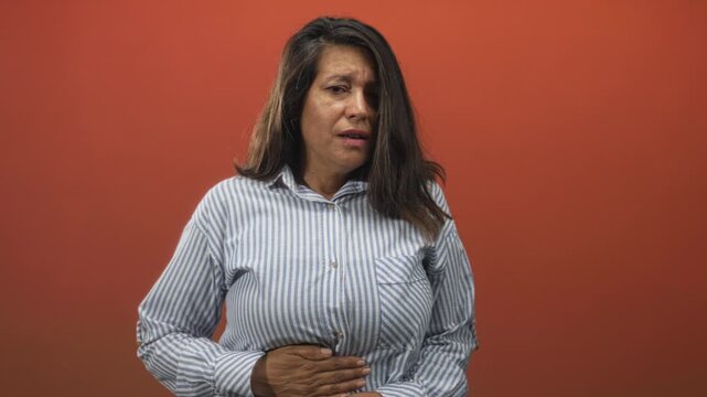 Woman middle aged brunette in striped shirt with hand on lower stomach and pained expression in studio with red wall; pain concern.
