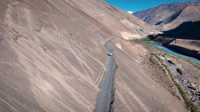 Cinematic drone tracking shot following a 4x4 SUV driving on a remote dirt road toward massive Himalayan mountains.