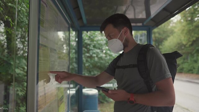 Male commuter wearing a protective mask studies bus schedule and route information at a bus stop in Trudering, Munich. Summer travel, public transport system and seasonal pollen allergy in the city.