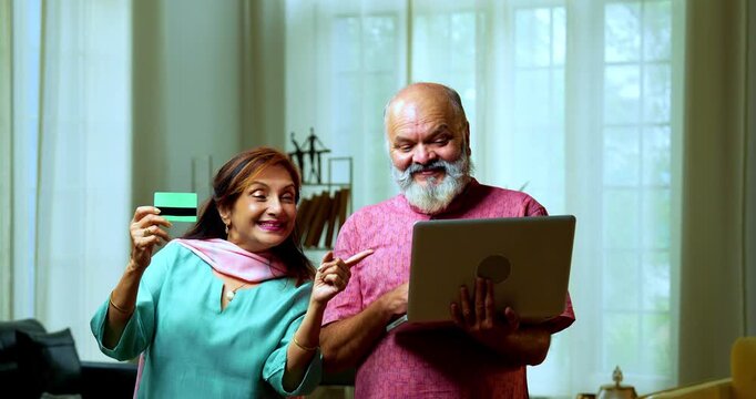 Senior Indian couple standing in a modern living room, using a debit card for an online transaction and showing it to the camera with confidence and smiles