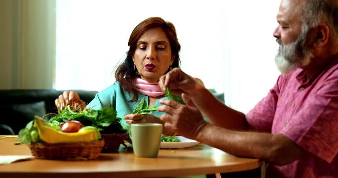 Senior Indian couple sitting together at a breakfast table and having a pleasant conversation while both are actively sorting fresh green vegetables in their kitche