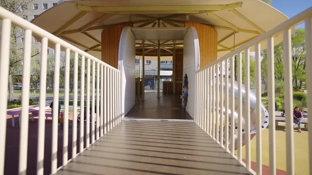 White bridge walkway leads toward round wooden tower. Railings frame approach route guiding room entry into elevated play structure