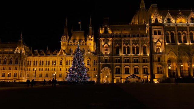 Real-time handheld 4K footage of the grand Budapest Parliament at night. Featuring golden architectural lighting and a festive Christmas tree on Kossuth Square in winter season.