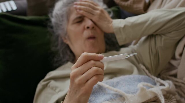 Medium selective focus shot of elderly Caucasian female seasonal flu sufferer lying on sofa covered with blanket, staring at thermometer with hand on forehead, coughing and sneezing