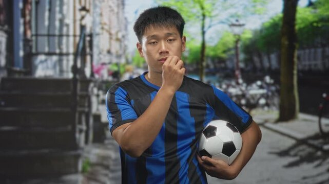Man in blue and black soccer jersey holding a soccer ball and pointing finger on street by bicycles and stone steps; determination.