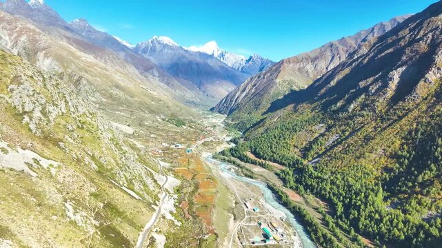 Cinematic drone pullback revealing a vast, colorful river valley and majestic snow-capped Himalayan peaks in Kinnaur.